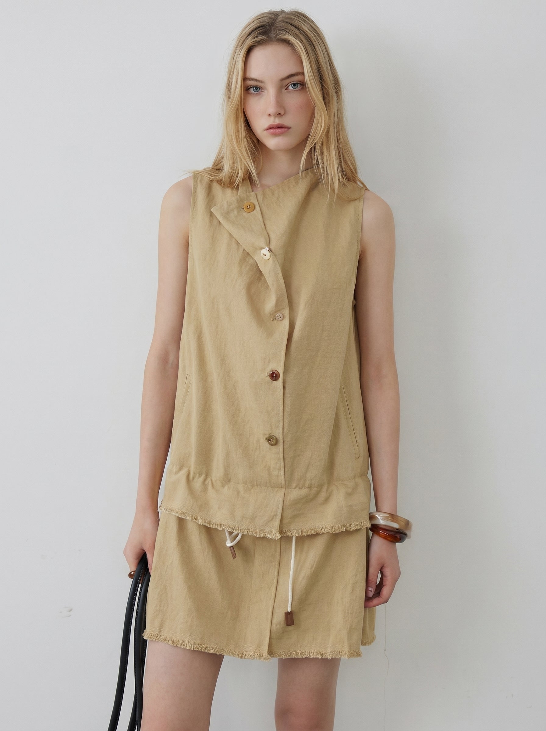 Front view of beige sleeveless mini dress featuring an asymmetric button front, drawstring waist, and raw hem details