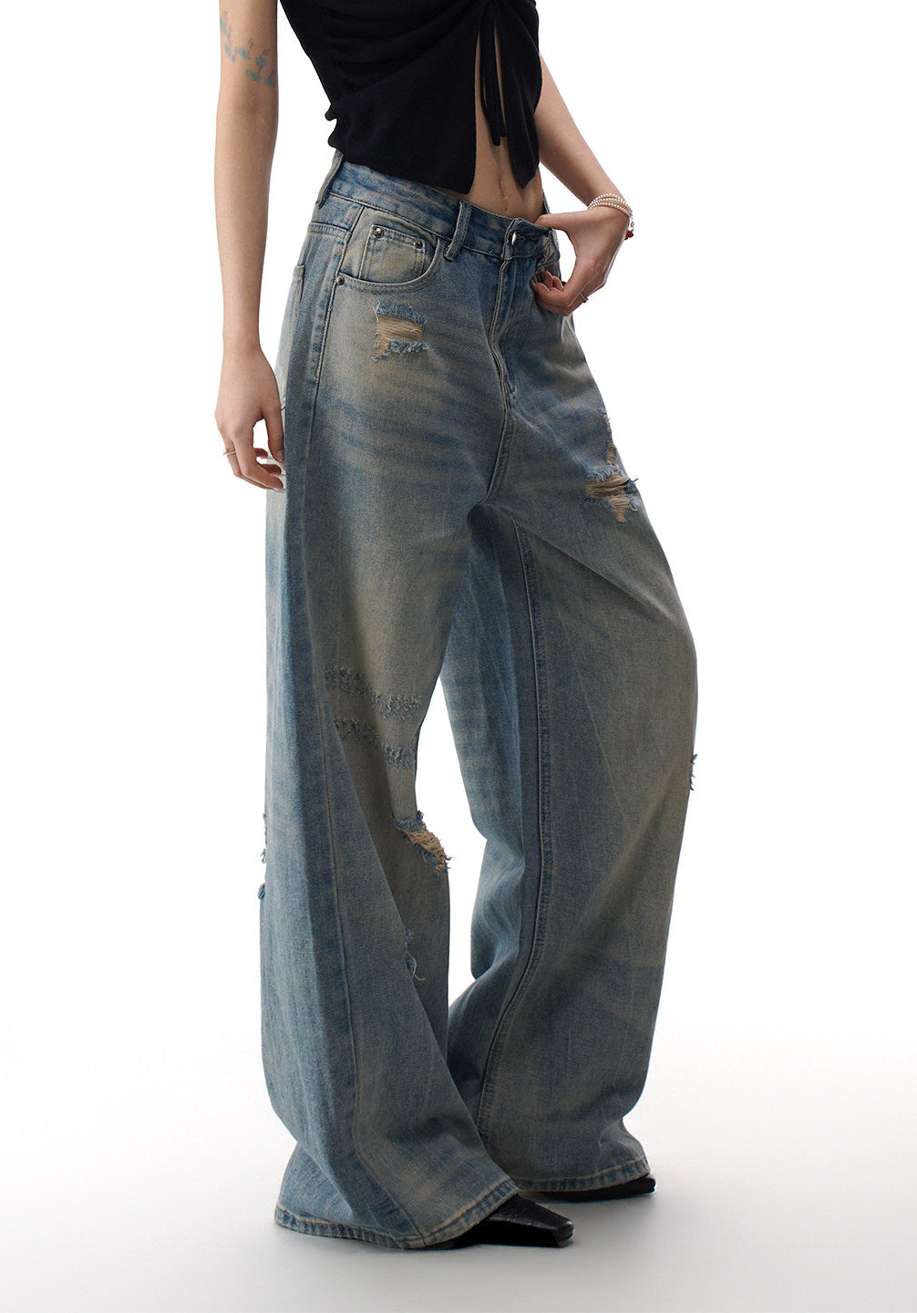 Close-up front detail of distressed blue wide leg jeans showing waist, pocket, button fly, faded wash, and ripped patches