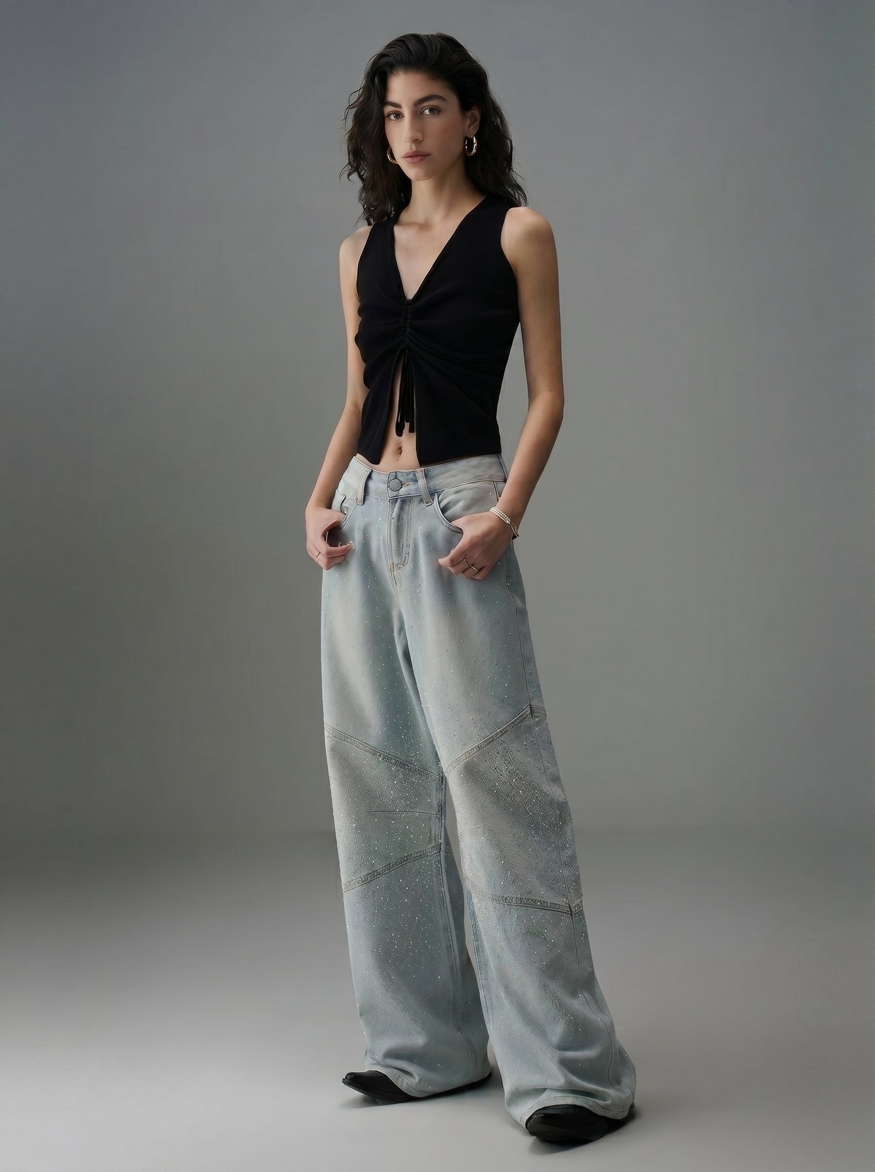 Model wearing light wash rhinestone wide leg jeans with a black ruched sleeveless top in a minimal studio setting