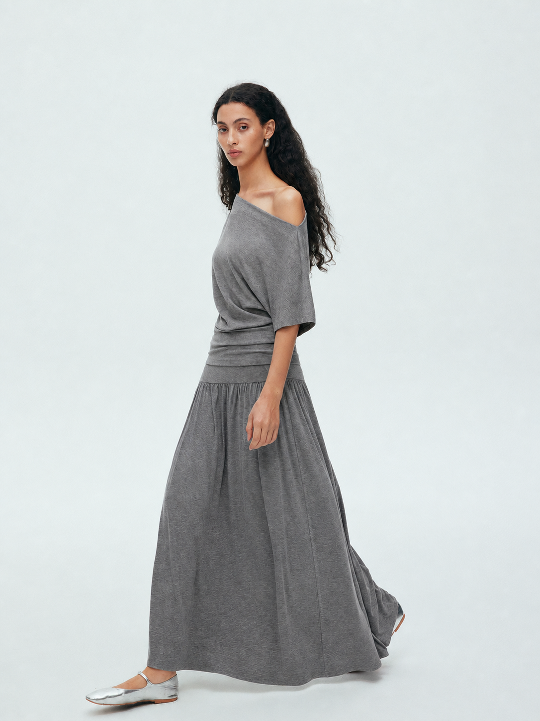 Model wearing a grey off shoulder draped top and matching maxi skirt set in a minimal studio setting