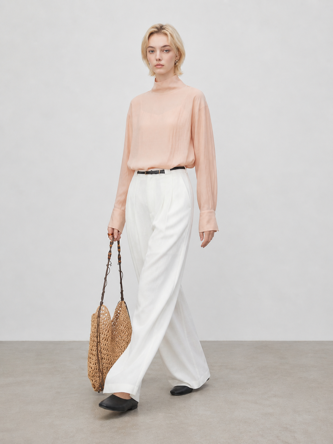 Model walking in peach sheer high neck blouse with white wide leg pants and woven tote bag