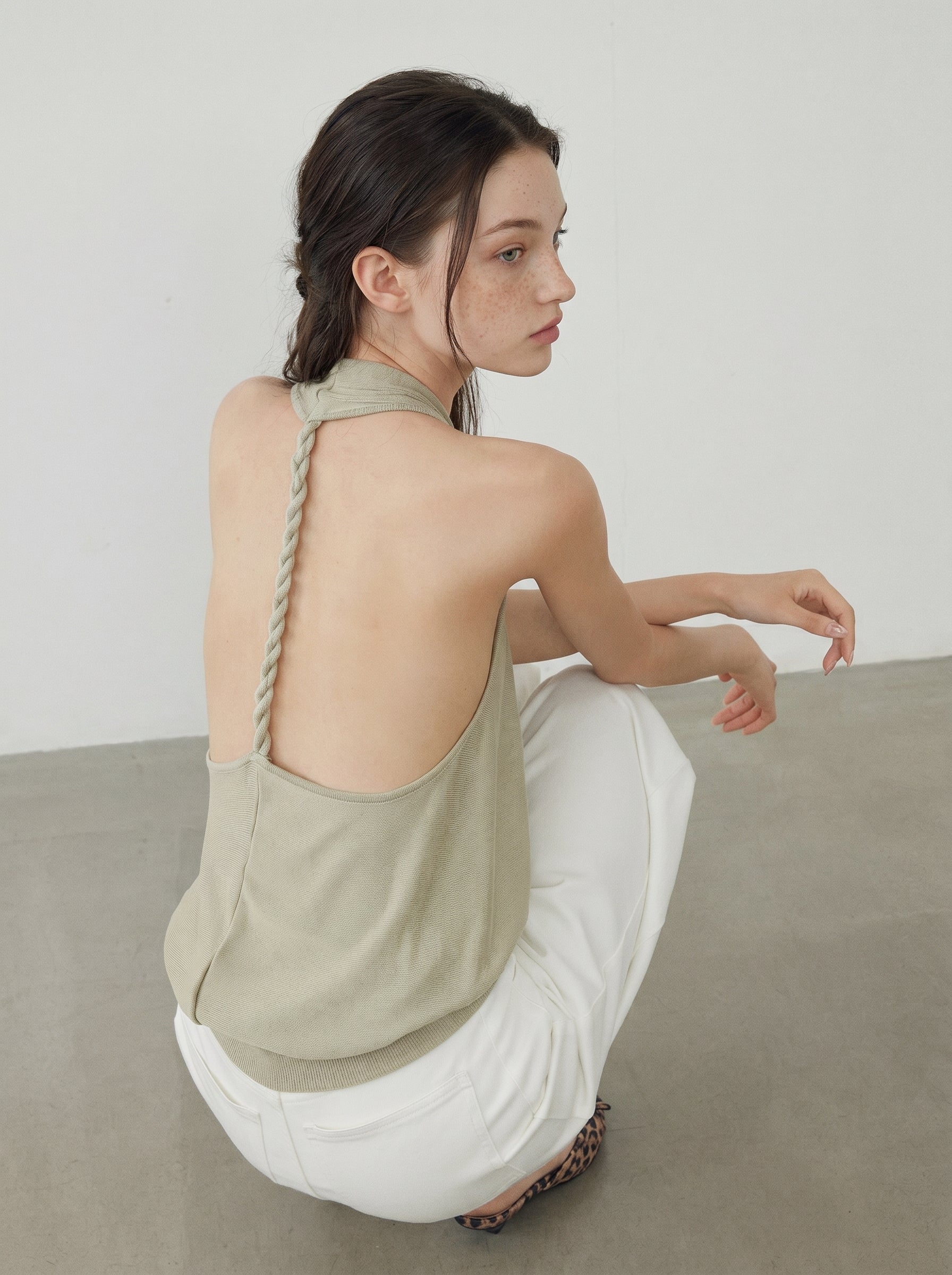 Model wearing a sage draped halter knit top with white wide leg pants in a relaxed seated pose